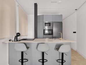 Eine moderne Küche mit Einbauofen, Mikrowelle und Kühlschrank. Es gibt drei Barhocker und eine Spüle mit Wasserhahn.