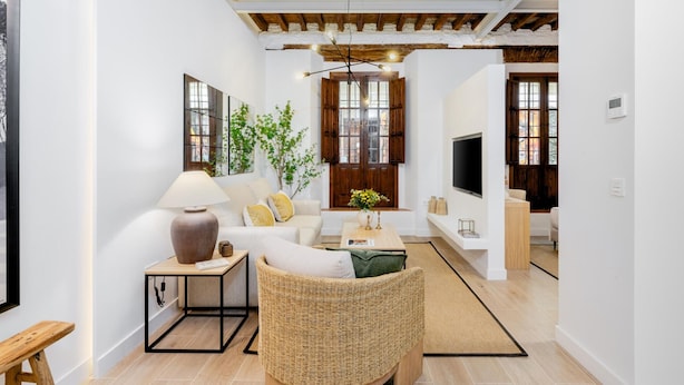 Una sala de estar moderna con un sofá, una mesa de centro y una silla de mimbre. Hay un televisor montado en la pared y una ventana con contraventanas de madera.