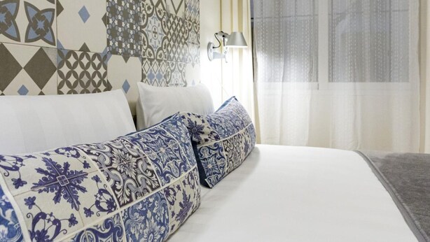 A hotel room with a patterned headboard, a bed with white bedding and blue and white patterned pillows, a bedside lamp, and a window with sheer curtains. - Charming Apt in the heart of Malaga with rooftop pool (Málaga)