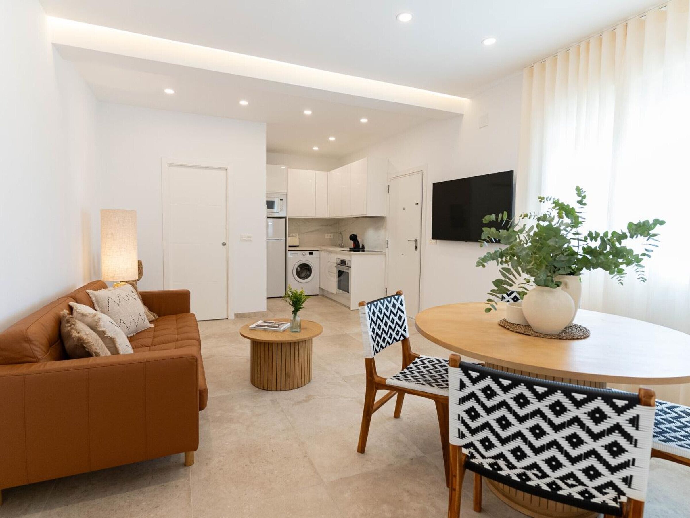 Una sala moderna con un sofá, sillas, una mesa de centro y una mesa de comedor. Se ve una cocina al fondo con electrodomésticos y armarios.