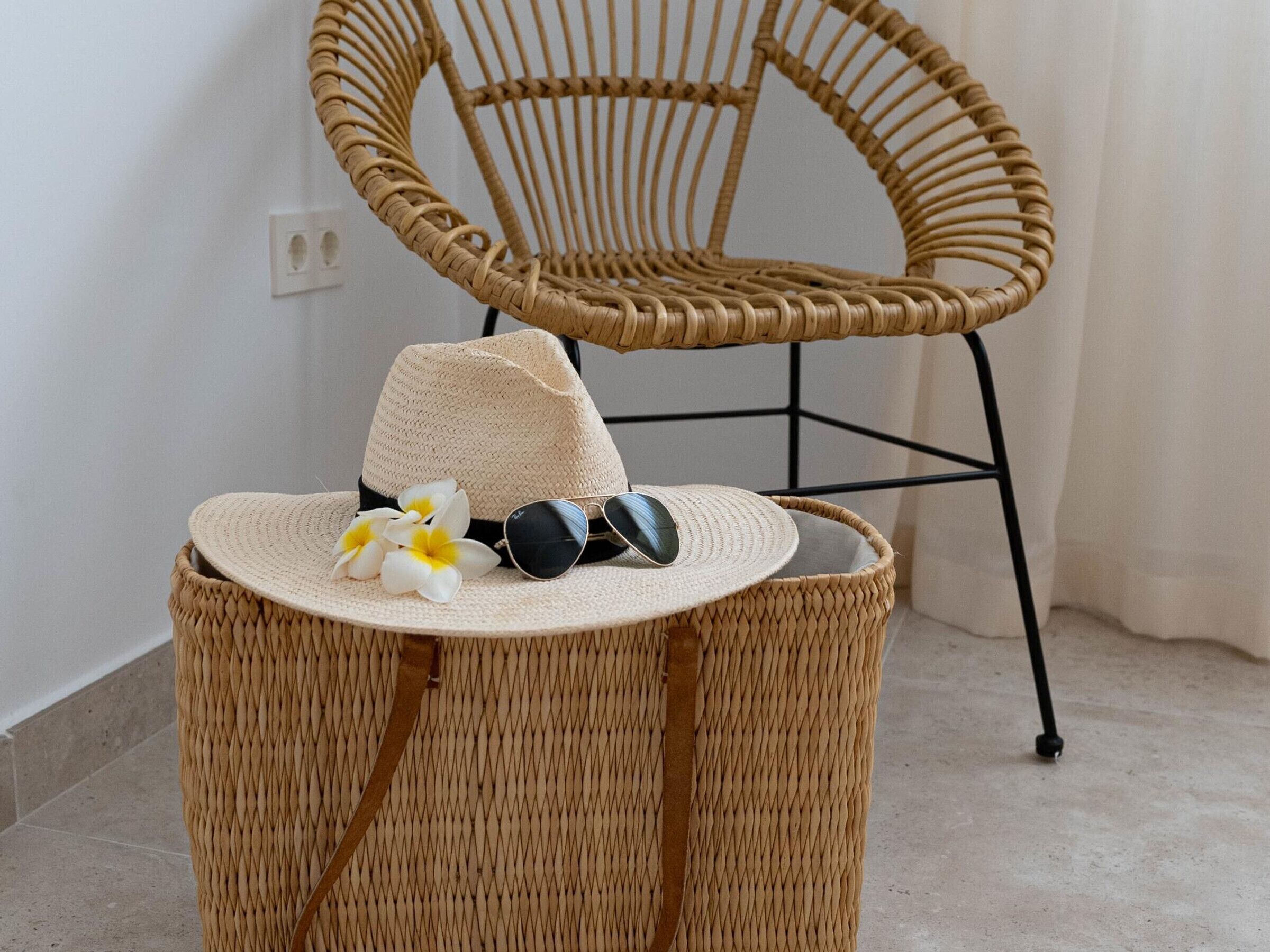 A wicker chair with a woven seat and backrest, a woven basket with a hat and sunglasses, and a white wall with a power outlet.