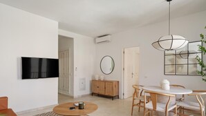 Una sala de estar moderna con televisor de pantalla plana, una mesa de centro redonda de madera, muebles de mimbre y un comedor con una mesa y sillas blancas.