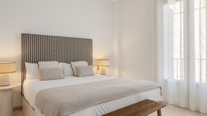 Un dormitorio con cama, lámparas de noche, un banco y una ventana con cortinas.