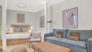 Een moderne woonkamer met een bank, een houten bijzettafeltje, een stoel en een bed met gekleurde kussens.