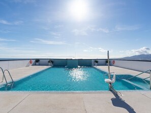 Une piscine à l’eau d’un bleu cristallin, entourée d’une terrasse en béton et de rampes métalliques, baignée par un soleil éclatant.