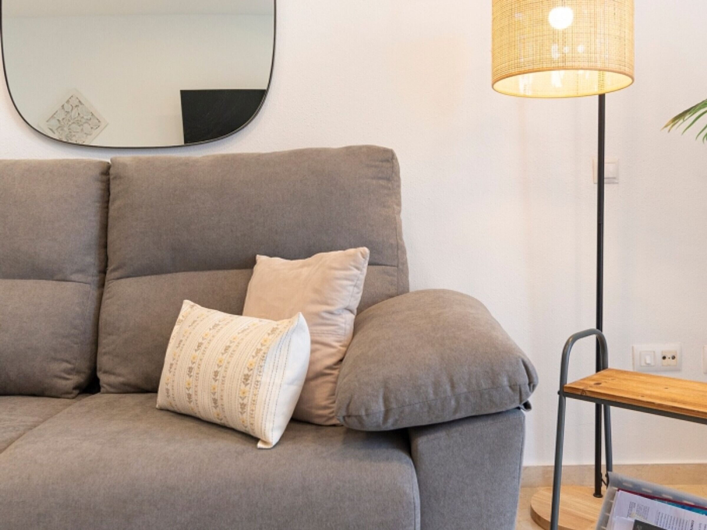 Un canapé gris agrémenté de coussins à motifs, un miroir rond accroché au mur et une lampe de sol avec un abat-jour en tissu tressé.
