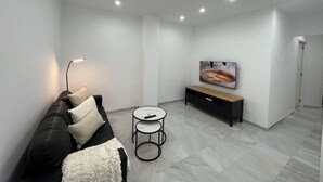 Una sala moderna con un sofá negro, un plaid blanco, una mesa de centro, un televisor en la pared y un mueble de madera.
