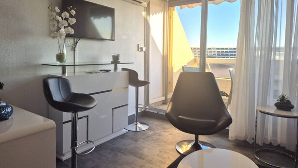 Een moderne hotelkamer met een balkon, een flatscreen televisie, een eettafel met glazen blad en twee zwarte barkrukken.