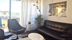 Een moderne woonkamer met een zwarte leren bank, een ronde salontafel en een zwarte stoel. Er staat een vloerlamp, een klein tafeltje met een plant en een groot raam met dunne gordijnen.
