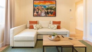 Ein Wohnzimmer mit einem weißen Sofa, zwei Zierkissen, einem Couchtisch aus Holz mit einer Pflanze und einem gerahmten Gemälde über dem Sofa.