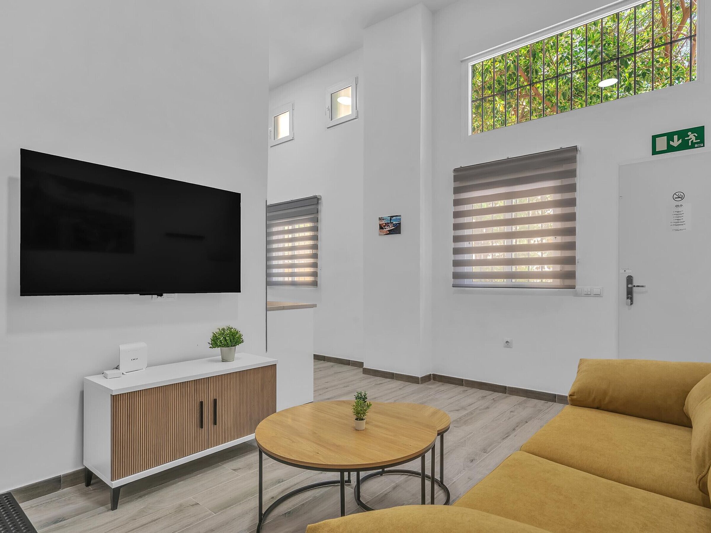Un área de espera moderna con una televisión de pantalla plana, un mueble de madera, una mesa redonda con una planta y un sofá amarillo.
