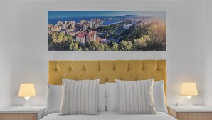 Quarto com vista para a cidade, cabeceira estofada, duas luminárias de cabeceira e cama com roupa de cama branca.