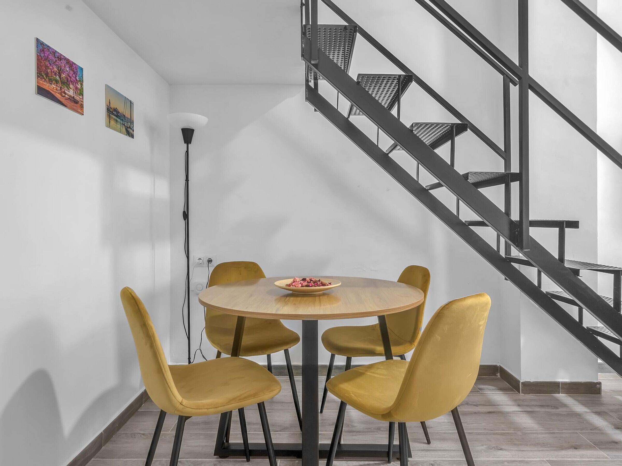Un comedor con una mesa redonda y cuatro sillas amarillas, una escalera con barandillas metálicas y dos cuadros colgados en la pared.