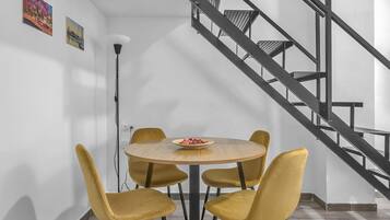 Un comedor con una mesa redonda y cuatro sillas amarillas, una escalera con barandillas metálicas y dos cuadros colgados en la pared.