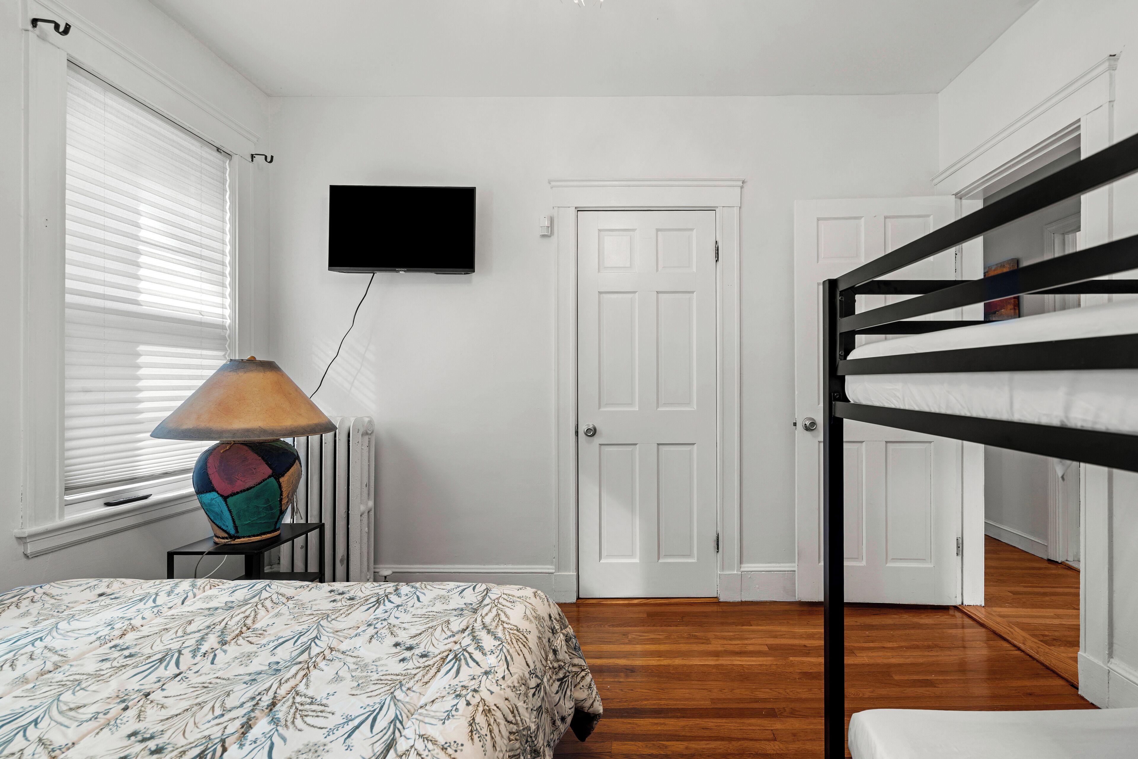 A bedroom with a bunk bed, a bed with a patterned bedspread, a wall-mounted TV, and a radiator.