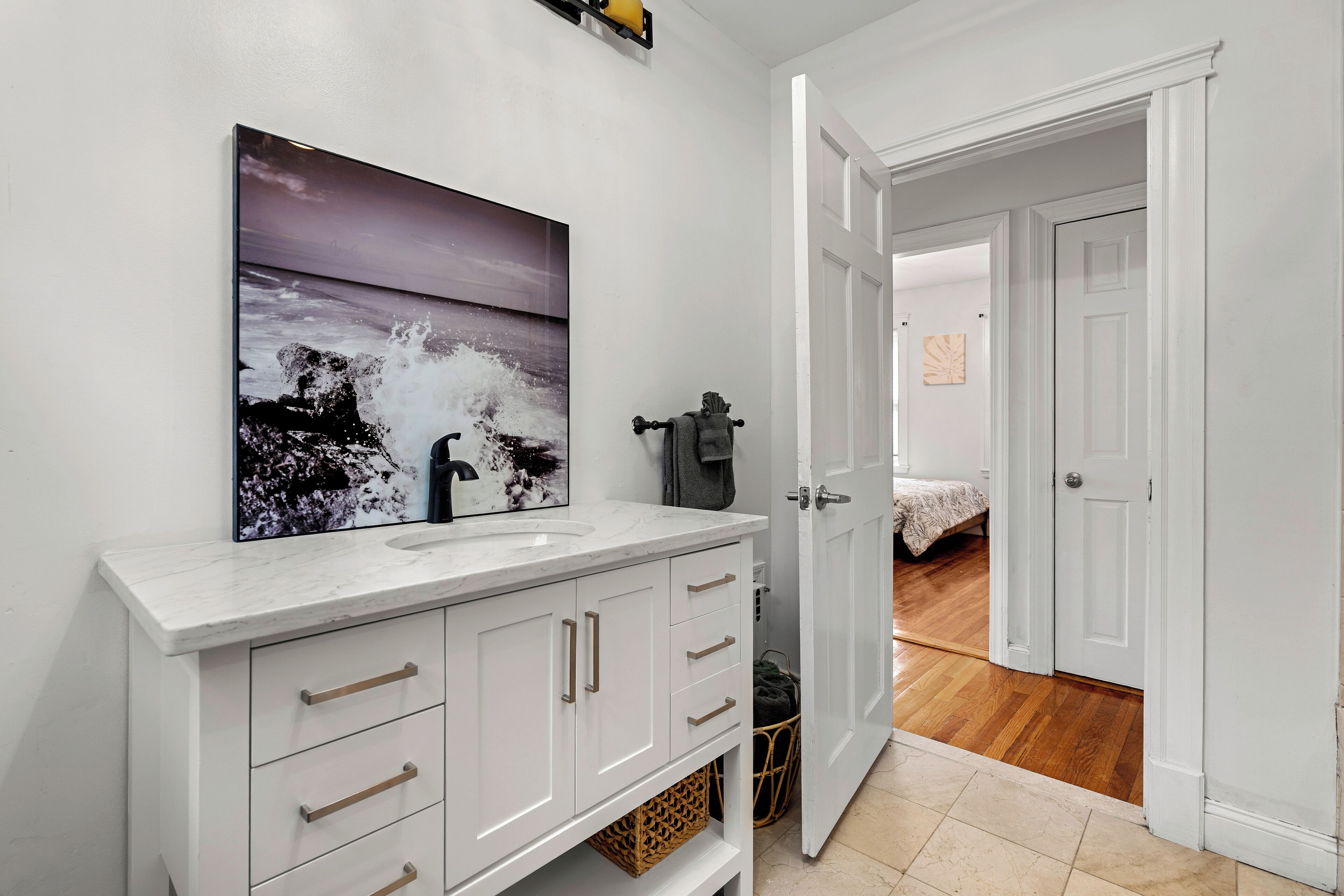 A bathroom with a white vanity, a large painting of a seascape, and a door leading to a bedroom with a bed and wooden flooring.