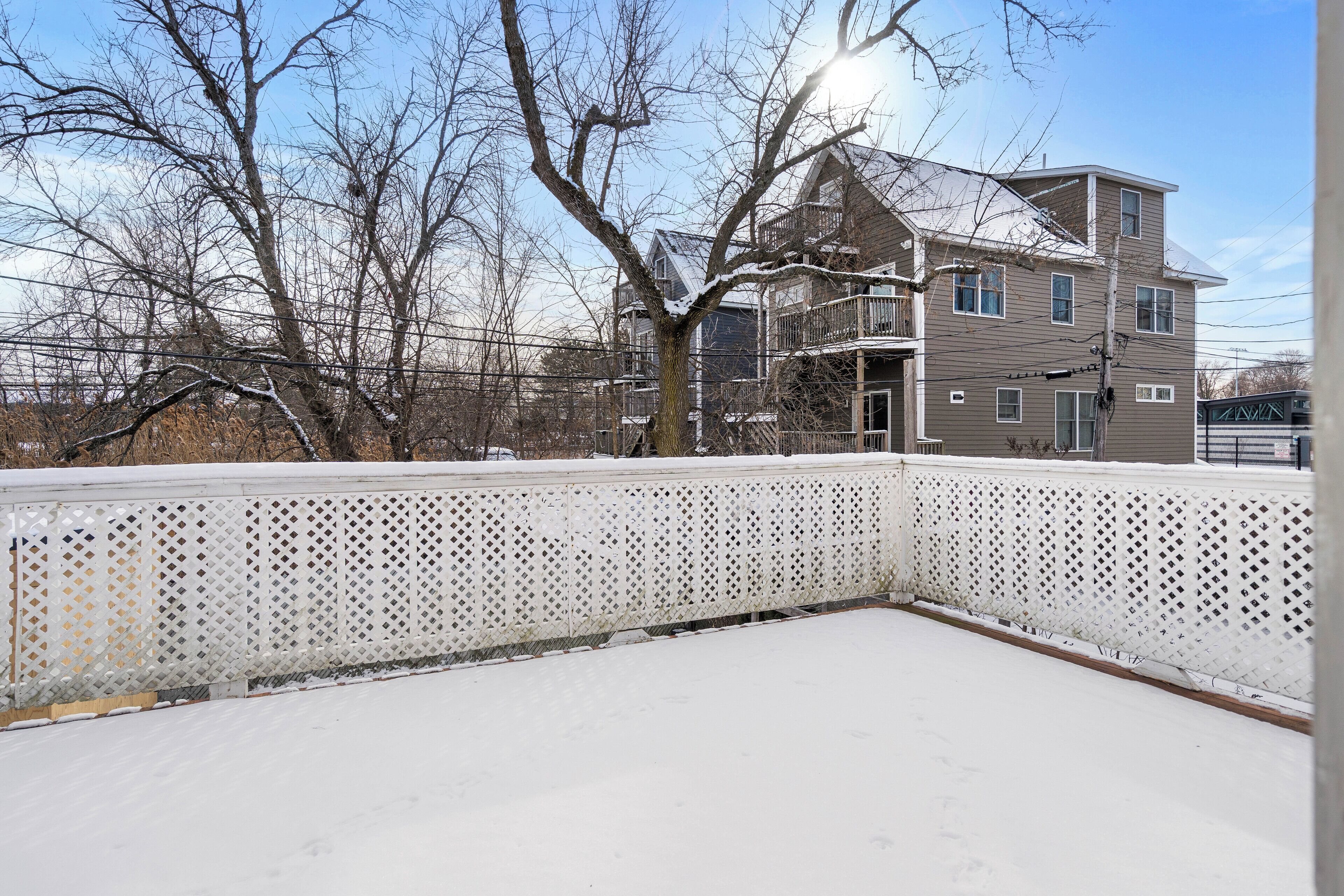 A snow-covered outdoor area with a white lattice fence, a multi-story residential building in the background, and bare trees.