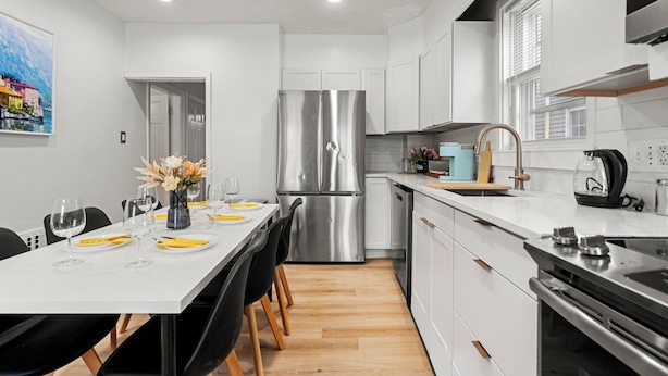 Une cuisine moderne avec des meubles blancs, un réfrigérateur en acier inoxydable et un coin repas avec une table dressée pour quatre personnes.