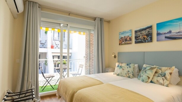 Uma cama de casal com almofadas florais, varanda com mesa e cadeiras, e uma pintura com vista para a cidade na parede.