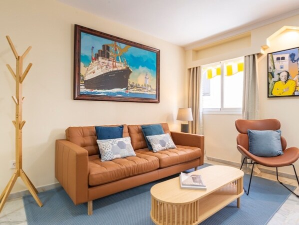 Ein Wohnzimmer mit einem braunen Ledersofa, einem Holztisch, einem blauen Teppich mit Muster, einem Holzständchen für Kleidung und einem Gemälde eines Schiffes an der Wand.