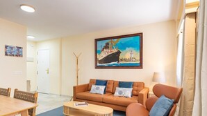 Sala de estar com sofá, mesa de centro e cadeiras. Há um quadro de um navio na parede.
