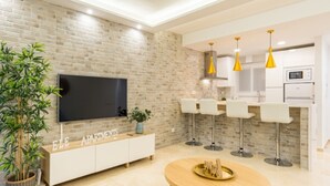 Ein modernes Wohnzimmer mit Flachbildfernseher an einer Ziegelwand, einem Holzespeisebereich und einer Küche mit weißen Schränken und gelben Pendelleuchten.