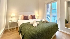 Un dormitorio moderno con una cama, dos mesitas de noche, una manta verde y vistas a un balcón.
