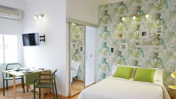 A bedroom with a bed, green pillows, a desk with chairs, a TV, and a wall with leaf-pattern wallpaper. - Modern Studio in Soho, Malaga (Málaga)