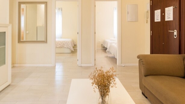 A hotel room with a large mirror, a beige sofa, a white coffee table, and a vase with dried flowers. There are two doors leading to a bedroom with a bed and a bathroom with a toilet and sink. - Bright 3-bedroom apartment in the heart of Málaga (Málaga)