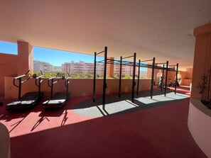 Un gimnasio en la azotea con equipo de ejercicio, incluyendo cintas de correr y una zona de dominadas, con vistas a un paisaje urbano.