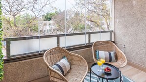 Un balcón con sillas de mimbre, una mesa pequeña con bebidas y vistas a árboles y edificios.