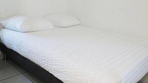 Cama bem arrumada com roupa de cama e almofadas brancas, cabeceira escura e chão de azulejos.