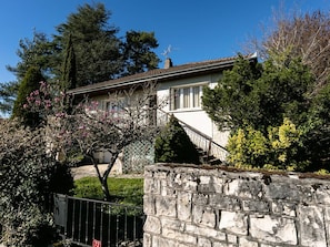 Un muro de piedra, una casa con escaleras y un árbol con flores rosadas.