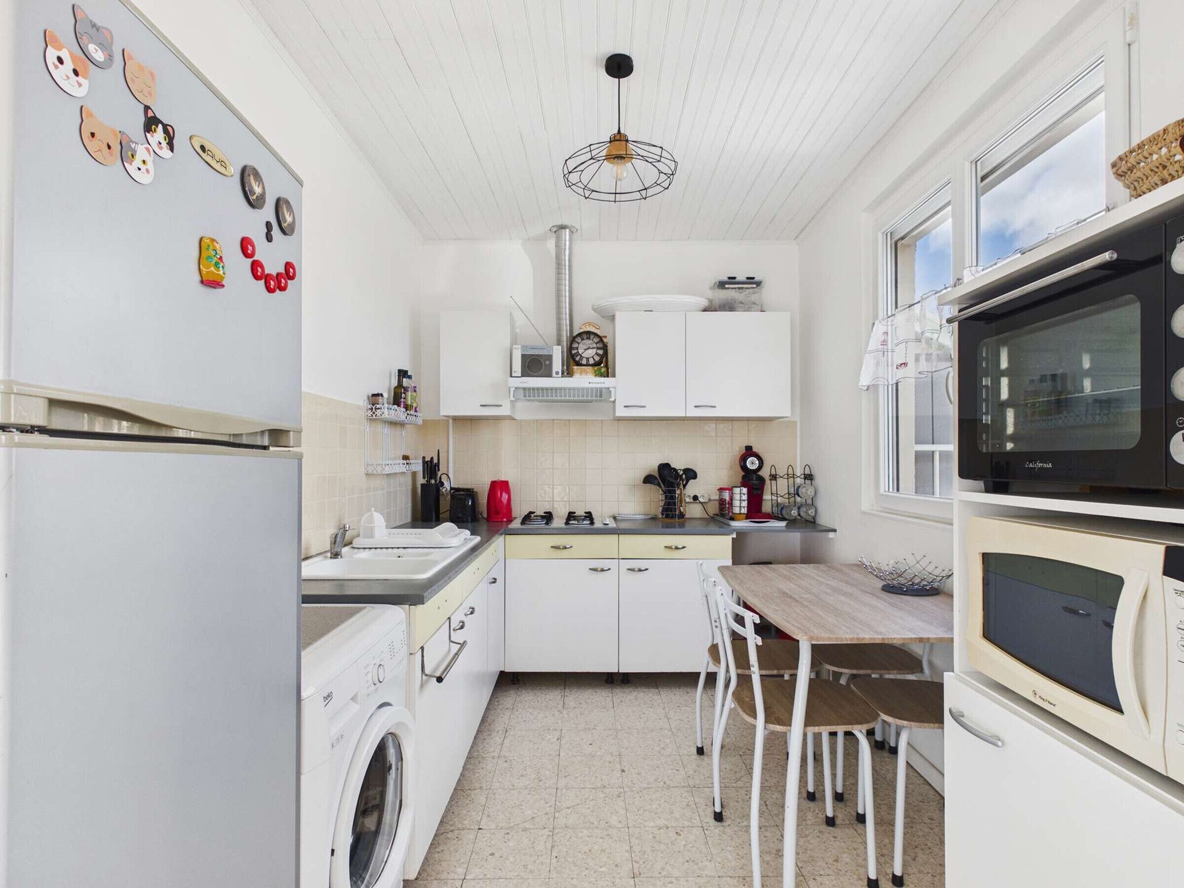 Een kleine keuken met een wasmachine, een eettafel met stoelen, een magnetron en een hangende lamp.
