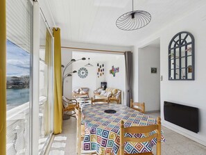 A room with a large window, a table with a colorful tablecloth, wicker furniture, and a unique ceiling light fixture. - Bright apartment with terrace, near beach and shops (Le Grau d'Agde)