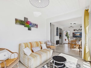 A living room with a striped sofa, a wicker chair, a dining area with a table and chairs, and a kitchen in the background. - Bright apartment with terrace, near beach and shops (Le Grau d'Agde)
