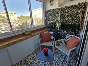 Uma pequena varanda com uma cadeira branca e uma mesa, um painel decorativo e vista para um parque de estacionamento e edifícios.