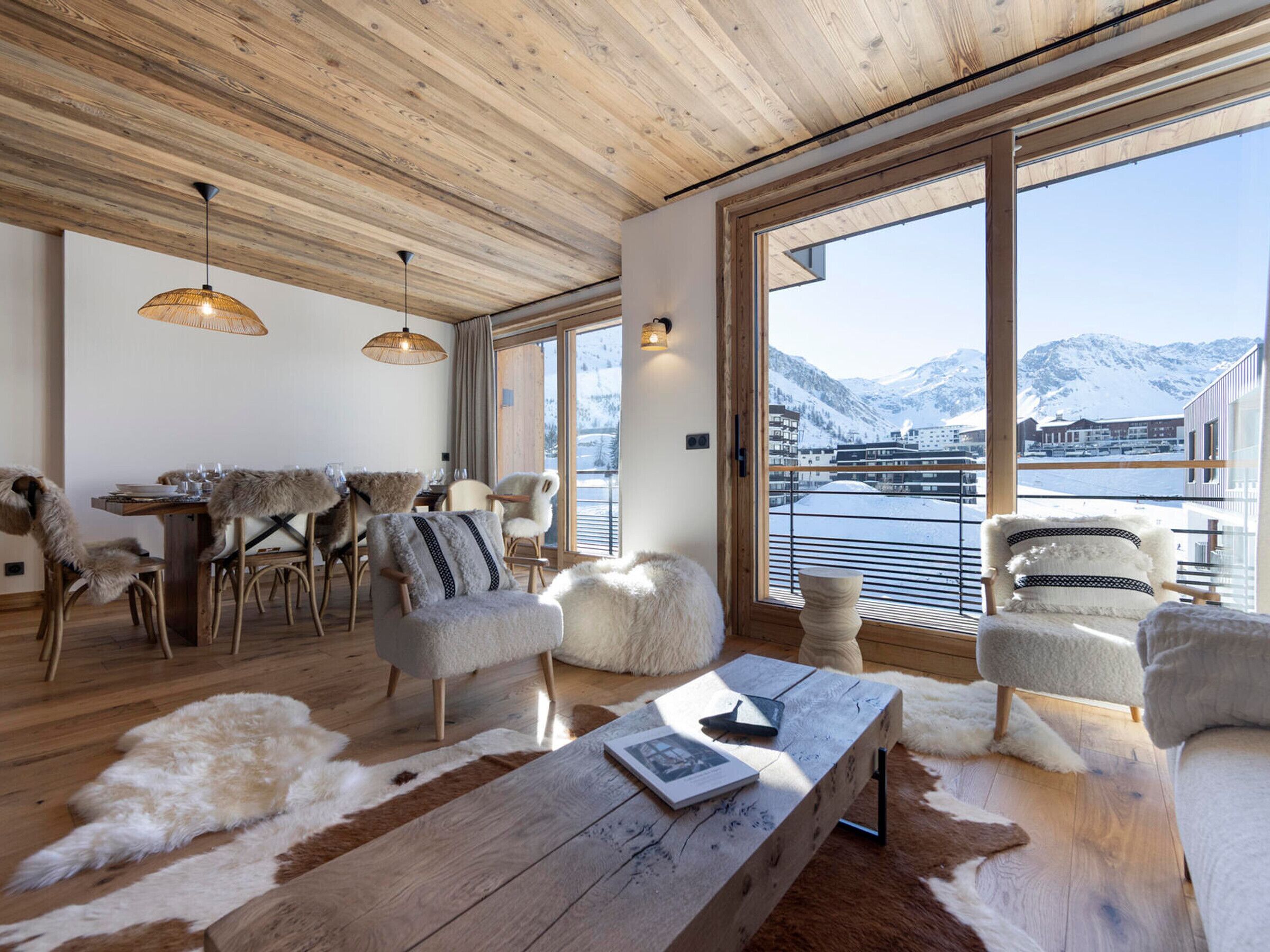 A spacious living area with a wooden ceiling, large windows with a mountain view, and a dining area with fur-covered chairs.