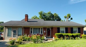 A single-story brick house with a gabled roof, a front porch, and a well-maintained front yard with various plants and a lawn.