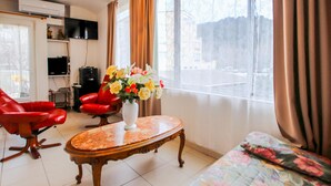 Una habitación con un sillón de cuero rojo, una mesa de centro de madera con un florero, un sofá con estampado floral, un televisor en la pared y una ventana con cortinas.