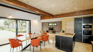 Een moderne keuken met een centraal kookeiland, een eethoek met oranje stoelen, en een televisie die aan een bakstenen muur is bevestigd.