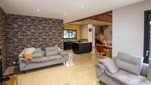Een moderne woonkamer met een accentmuur van baksteen, een grijze bank en een vloer van beige tegels.