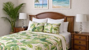 Ein Schlafzimmer mit einem Bett, zwei Nachttischen, einem Holzkopfende, einer großen Topfpflanze und zwei Wandbildern mit tropischen Motiven.