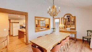 Un comedor espacioso con una mesa de madera y sillas a juego, una araña y una cocina visible a través de un vano abierto.