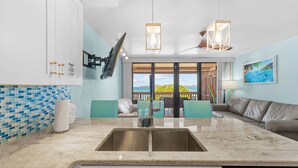 A modern kitchen with a large island, a dining area with teal chairs, and a view of the ocean through the sliding doors. - Oceanfront, Sapphire Beach, Pool, 2 bedrooms, 2 bath (St. Thomas)