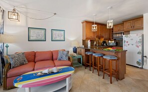 A compact living space with a sofa, a colorful coffee table, a kitchen area with wooden cabinets, a refrigerator with floral design, and a dining area with stools. - Sapphire Beach, Oceanfront, Pool, Steps to beach, Sleeps 4 (St. Thomas)