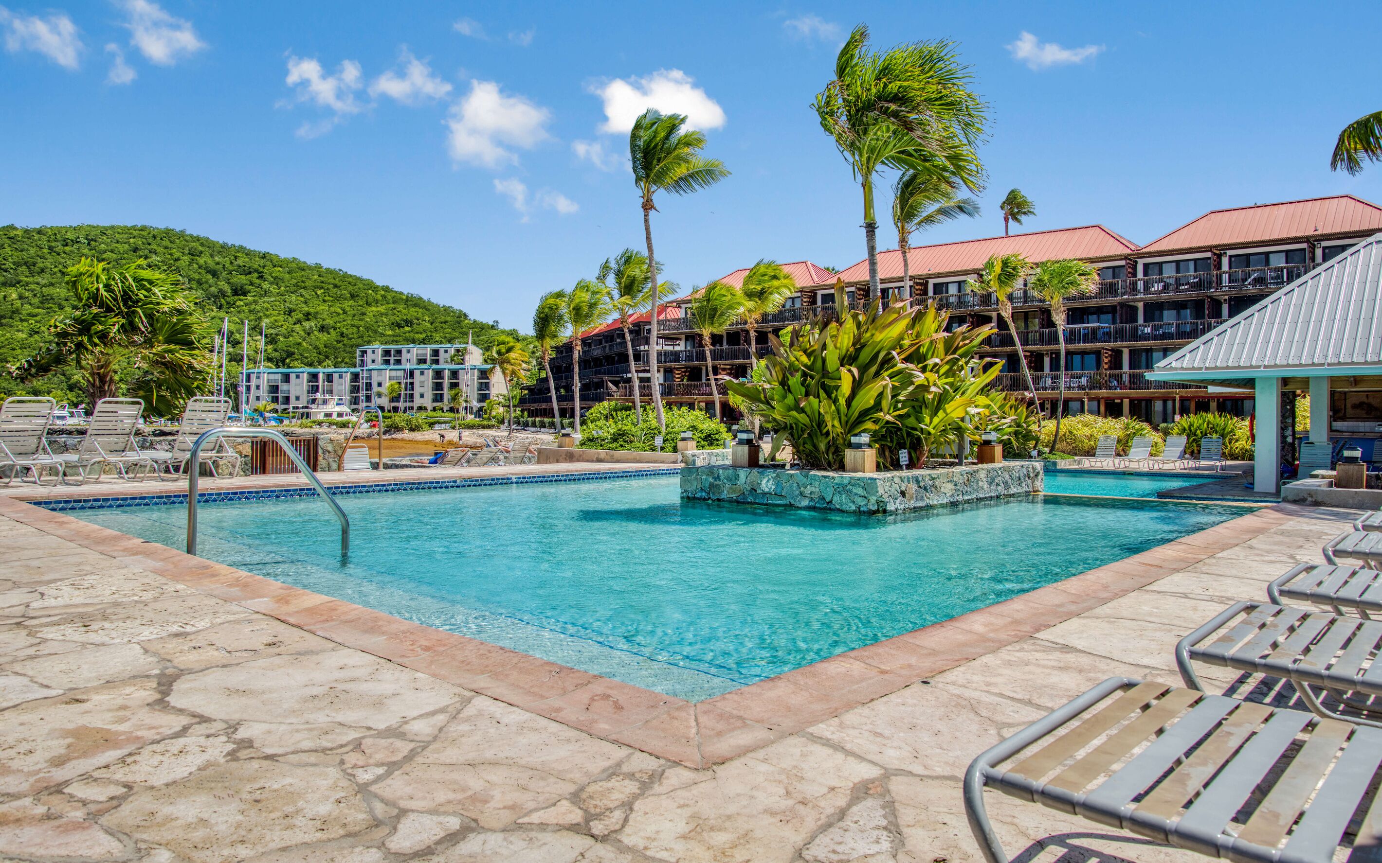 Zona de piscina en un resort con múltiples sillas de descanso, una piscina azul cristalina y un fondo de edificios de hotel y palmeras.