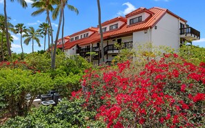 Un hôtel à plusieurs étages, avec un toit rouge, entouré d’une végétation luxuriante et de fleurs rouges.