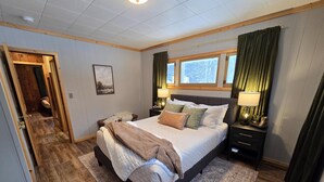 Un dormitorio con una cama grande, dos mesitas de noche, una ventana con cortinas, suelo de madera y una puerta que da a otra habitación.