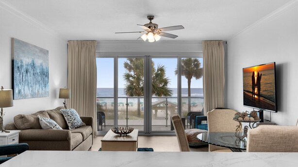 Ein geräumiges Wohnzimmer mit einem großen Fenster und Blick auf den Strand und Palmen. Es gibt einen Deckenventilator, einen an der Wand befestigten Flachbildfernseher sowie einen Essbereich mit Tisch und Stühlen.